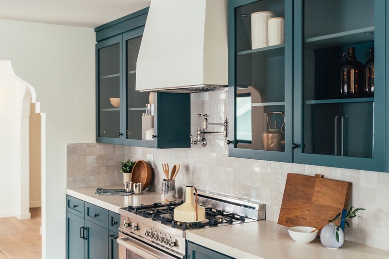 Kitchen stove with white hood, ceramic square tile, cream countertops, and blue green painted cabinets. Open cabinetry on top.