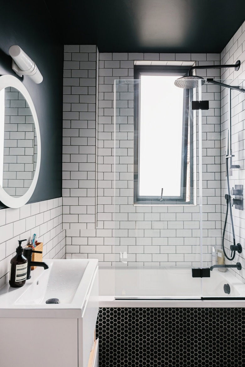 White-tiled bathroom with black ceiling, silver shower head, bathtub, and white sink with black faucet