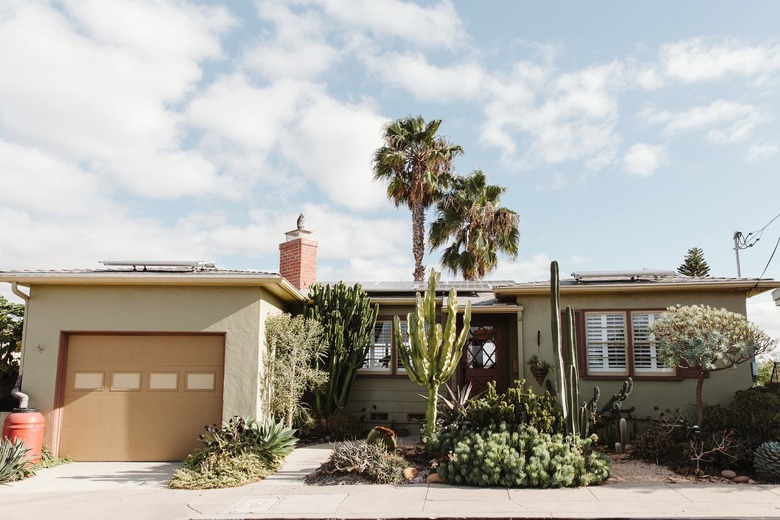 front of light green house with brown garage door, chimney, and greenery in front yard