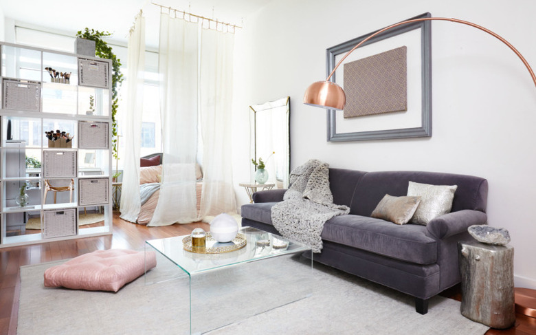 studio bedroom idea with a gray couch, clear acrylic coffee table, tall bookcase divider, and a bed behind sheer white curtains