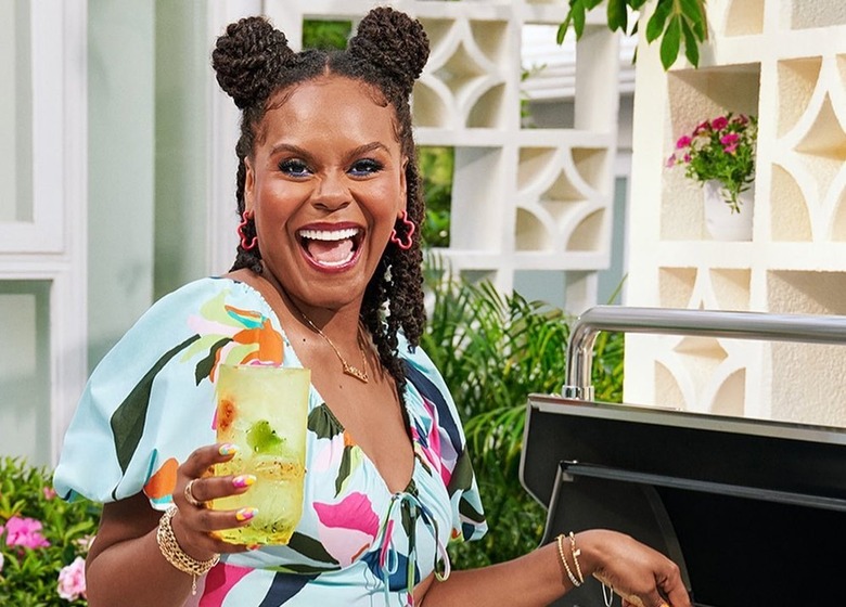 Tabitha Brown standing a grill, holding a drink in a yellow glass