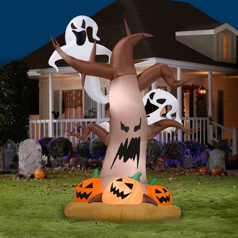 A blow-up shaped like an evil tree with a scary face. Two white ghosts and coming out from behind the tree and three jack-o'-lanterns are at the base. Behind the inflatable, there is a white house with a wraparound porch. On the lawn, there are also assorted Halloween decorations like gravestone, spiderwebs, and small orange pumpkins.