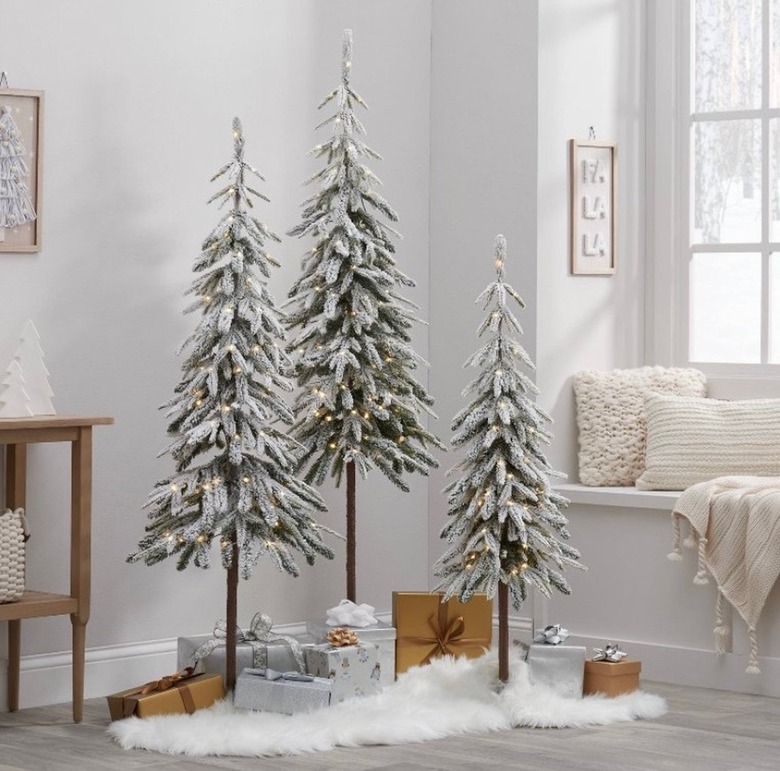 Three downswept flocked alpine balsam artificial Christmas trees. The three trees are lit, with faux snow underneath them with an array of brown and silver wrapped presents. There is a window and a window seat to the right, with two cream colored pillows and a decorative wall print that reads 