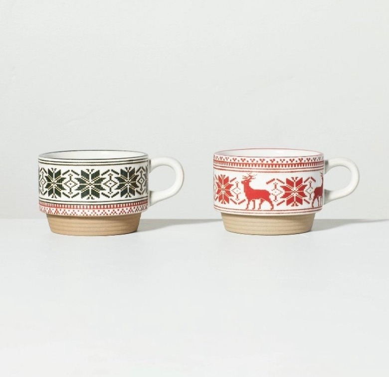 Two small mugs, both with tan bottoms and white handles, the one on the left is green snowflake Fair Isle print, and the one on the right is red reindeer and snowflake Fair Isle print