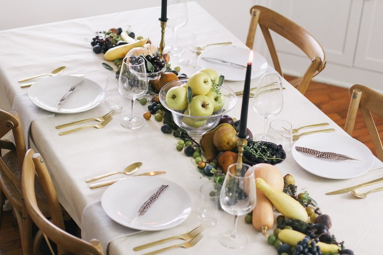 a line of produce, including grapes, pears, cherries, and squash runs down the length of a table with a white cloth and gold cutlery