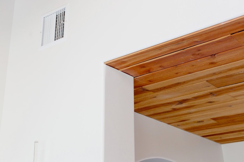 A white air vent on a white wall near the ceiling. Through an open doorway, a wood-paneled area of the ceiling.