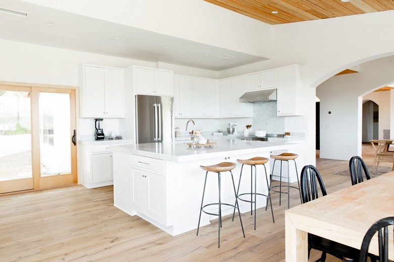 Open kitchen area with a white kitchen island. Three bar stools with wooden seats and metal legs in front of the island. Behind the island, there's a stainless steel refrigerator and white cabinets run along the wall. Next to the kitchen, sliding glass doors. In the foreground, a wooden dining table surrounded by black winsdor chairs.