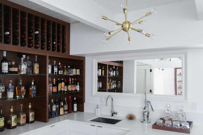 Built-in wood cabinets with bottles of wine and liquor, and a gold modernist bulb chandelier, over a sink.