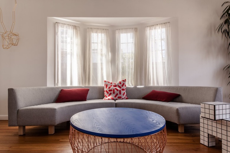 Curved gray sofa with red accent pillows. Round coffee table with a black wood top and intricate steel wire base. A multi-cubed side table.