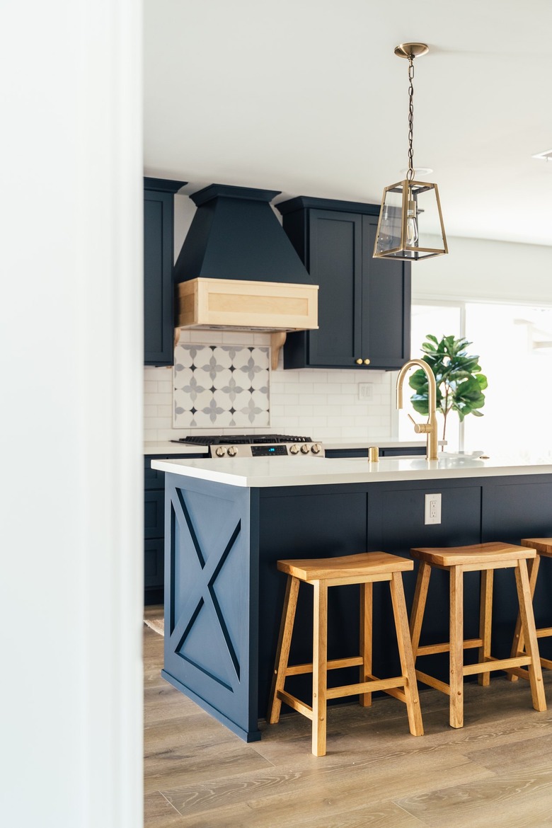 Dark blue kitchen cabinets, ornate backsplash, kitchen island with wood stools, gold lantern pendant light.