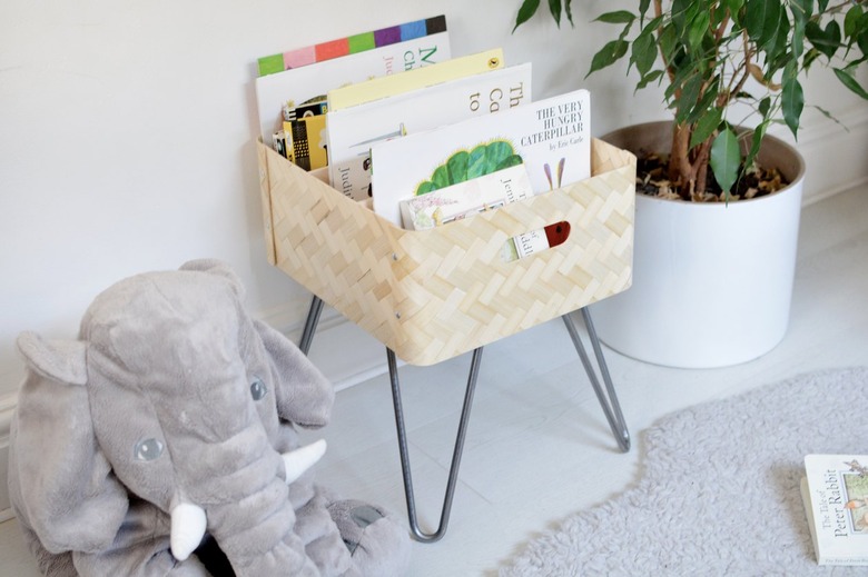 Light tan book bin with metal legs and picture books in a white room with grey rug next to elephant stuffed animal and medium-sized plant