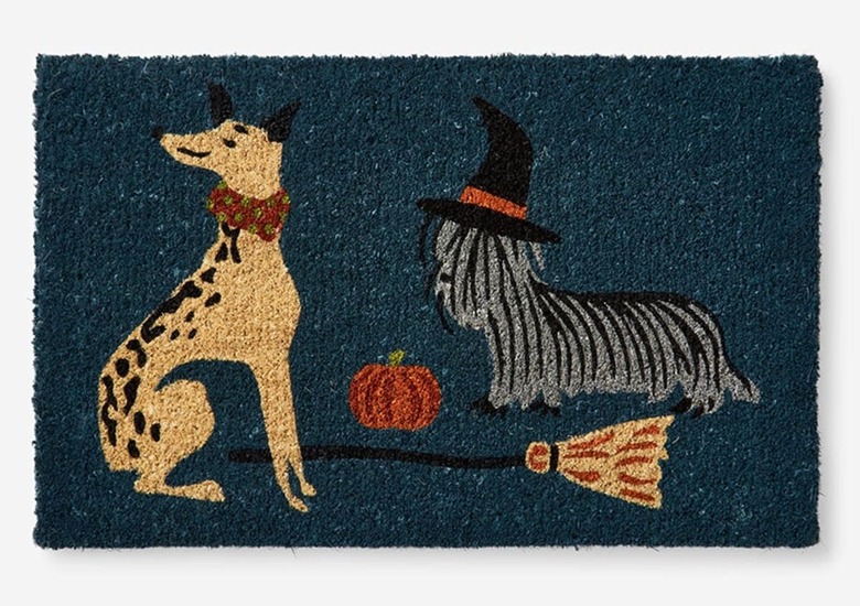 A navy blue doormat featuring two dogs. One is wearing a witch's hat and in between them, there is a broom and small orange pumpkin.