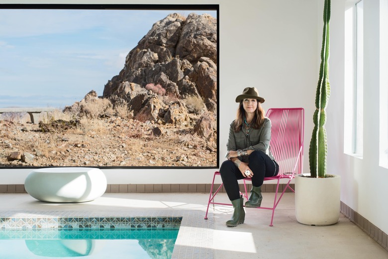 Kelly Van Patter sits by the inside pool on a pink mid-century style lounge chair by a large slider window with desert view.