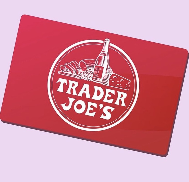 A red Trader Joe's gift card against a pink background. The Trader Joe's logo is in the middle of the card with white text and a picture of sliced bread, grapes, a bottle of wine and cheese, also in white.