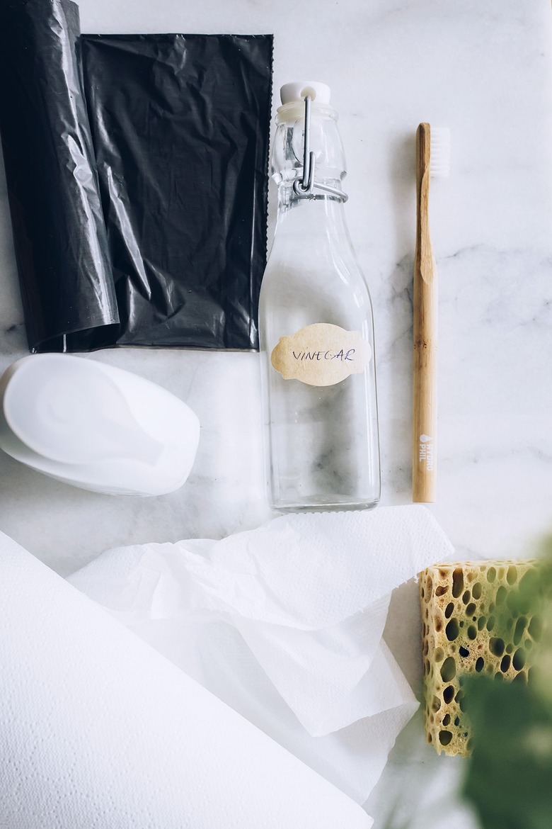 Black garbage bag, soap, empty glass bottle, wood toothbrush, paper towels, and sponger on white countertop