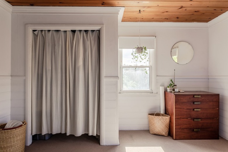 A sunlit bedroom with a curtain divider, hanging plant, wood dresser, round mirror, and baskets.