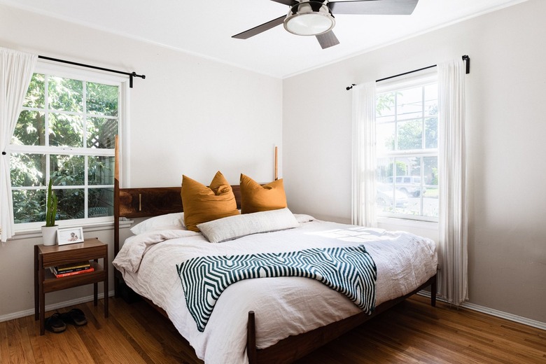 Wood headboard with white bedding, yellow pillows, blue-white lined pattern blanket. Wood night table and white curtains.