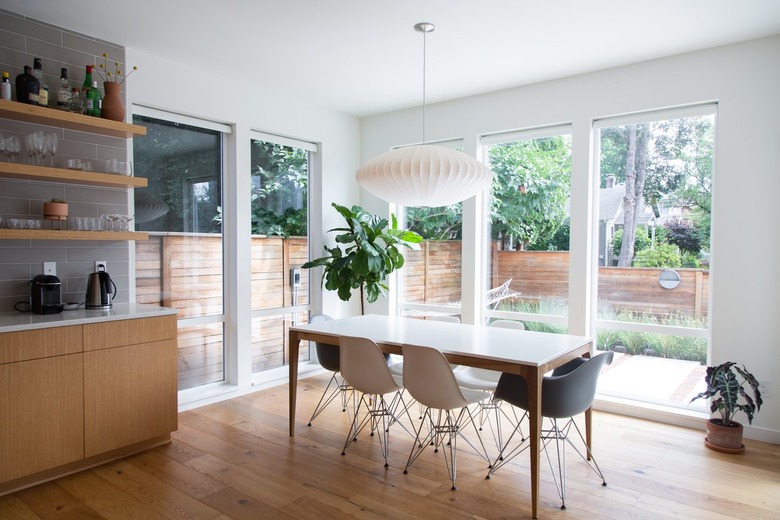 Dining room with picture windows, white oval pendant light, white dining table, Modernist chairs. Wood cabinets, shelves, gray wall tiles.