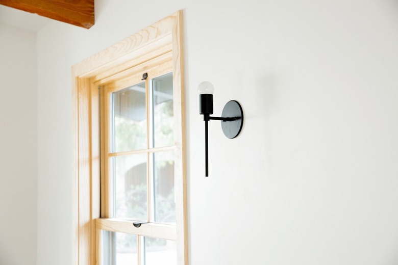 A window with a light wood frame against a white wall, near the corner of the room. A black metal scone is next to the window.