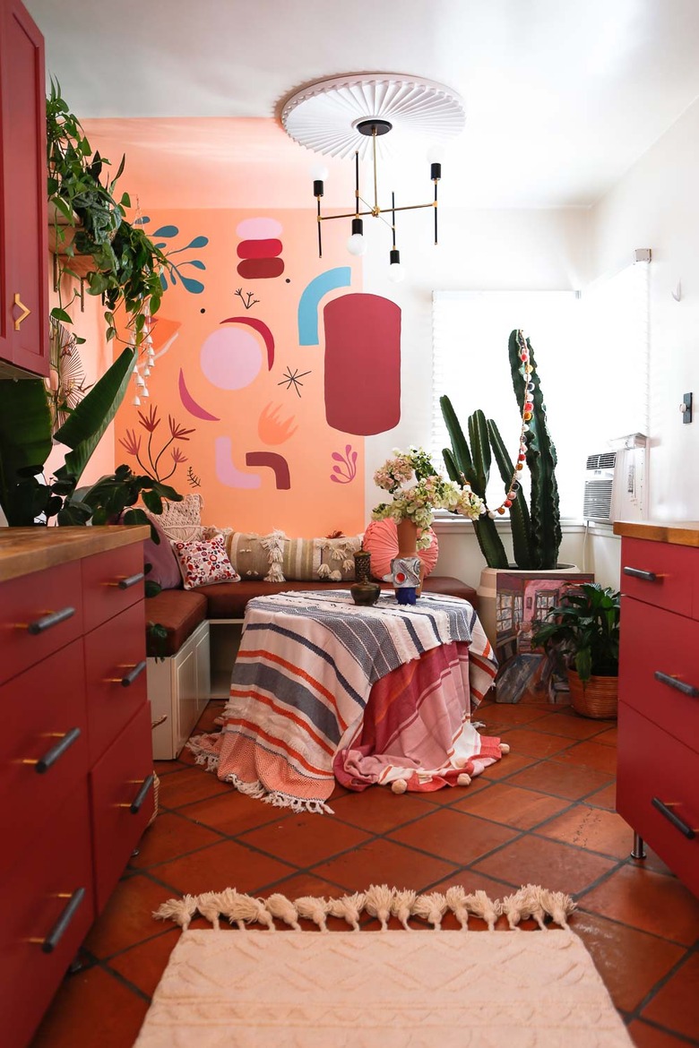 vermillion color kitchen cabinets, terracotta tile floor, art painted on wall, corner built in dining space, textiles over dining table, modern chandelier, plants.