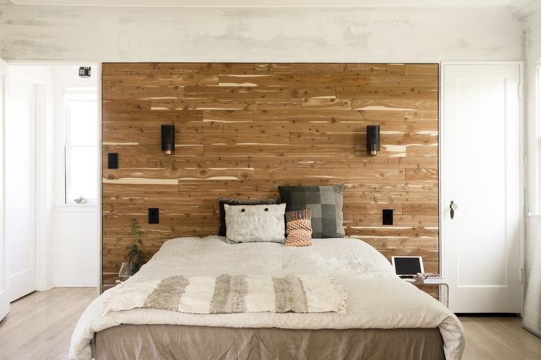 A bed with multi-colored neutral bedding and pillows. A wood accent wall behind with sconces.