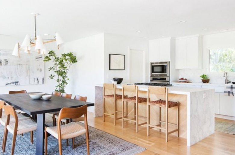 Dining room with open kitchen featuring marble waterfall kitchen island, cooktop, barstools and hardwood floors.