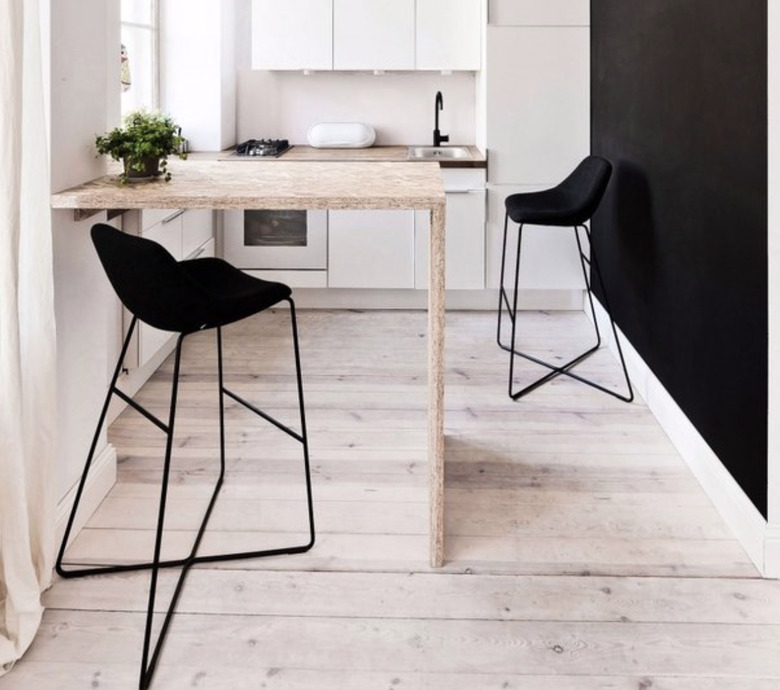 Small waterfall kitchen island in white kitchen with light wood floors, black accent wall and black barstools