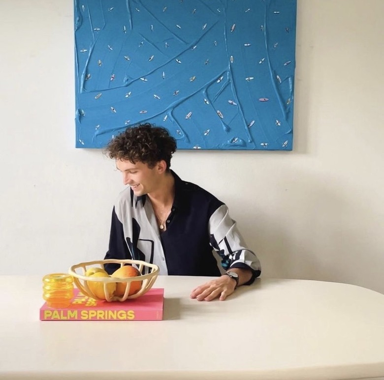 Artist Werner Bronkhorst sits at a white kitchen table and is looking to the side and smiling. He is a white man with curly short brown hair and is wearing a white and gray long-sleeve button-up top. A bright pink book with 