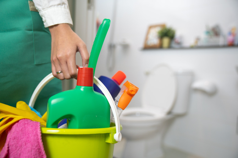 Woman standing bathroom holding a yellow plastic bucket full of bathroom cleaning supplies.