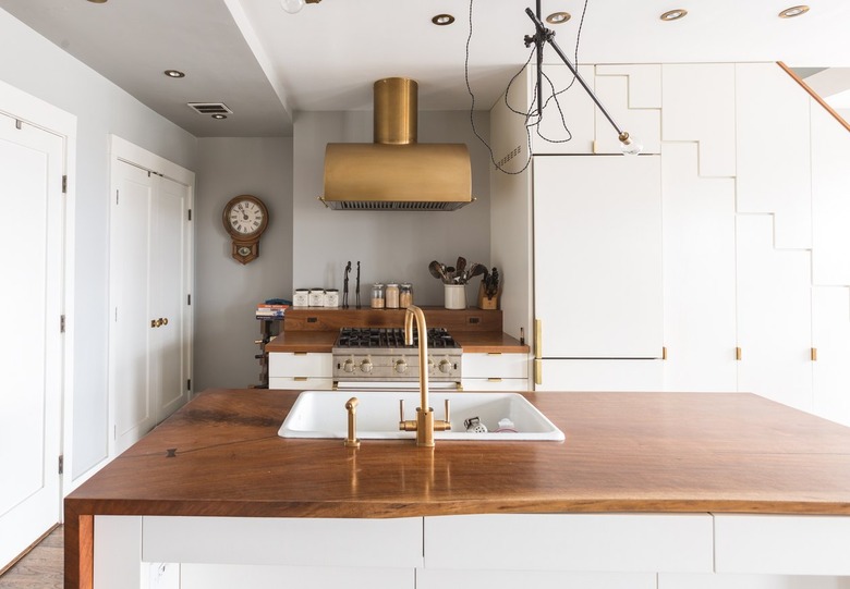 A white kitchen island with a wood counter and brass faucet sink. A brass hood over a stovetop and wood counter.