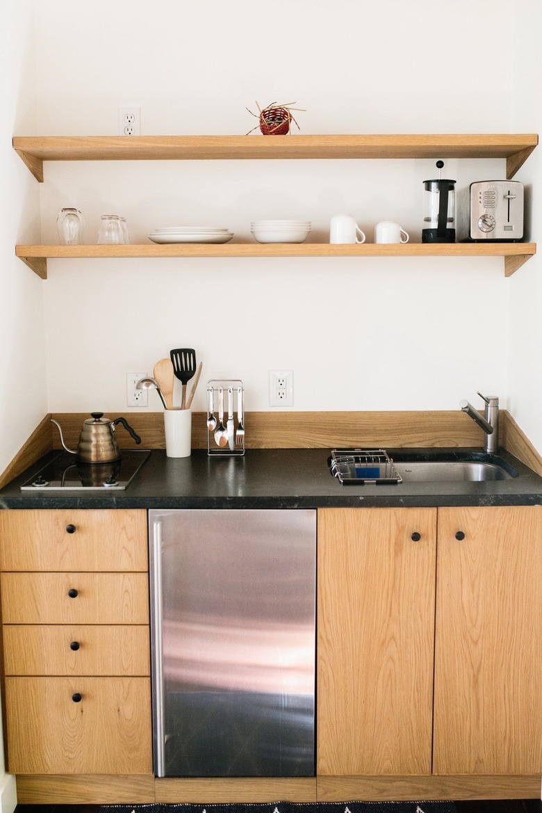 A small kitchen with light wood cabinets, black counters, and two shelves. A metal sink and small stovetop are on either end.