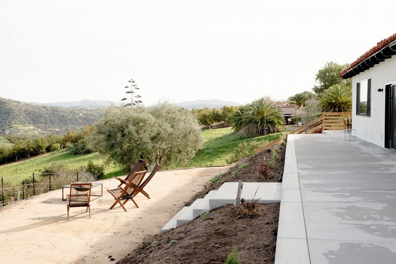 Concrete walkway with steps leading down to a dirt patio area where there are four wooden lounge chairs and a fire pit. The seating area overlooks rolling hills and a young vineyard.