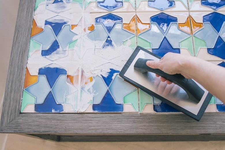 Person using a square notch trowel tile and grout, to secure orange and blue deco tiles to a wood coffee tabletop