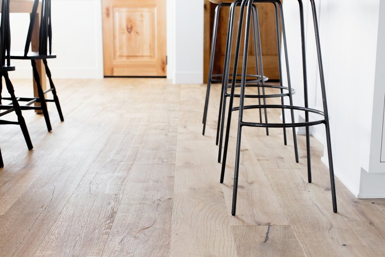 Chairs between kitchen and dining area with a pathway between leading to a wooden door. The floor is wood and the walls are white. On the right, three bar stools with metal legs. On the left, black Windsor chairs.