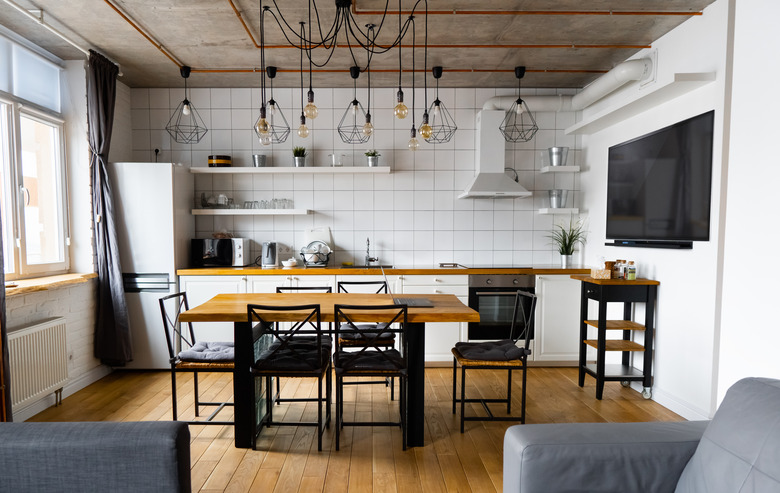 Modern scandinavian an eat-in kitchen interior design with big wooden table and chairs against light wood floor, bright white walls and furnitures with TV, appliances and hanging light bulbs