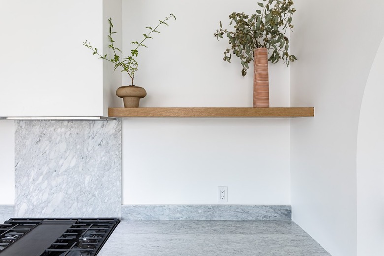 Shelving unit with two small plants over marble backsplash above black stovetop and marble countertop against white wall