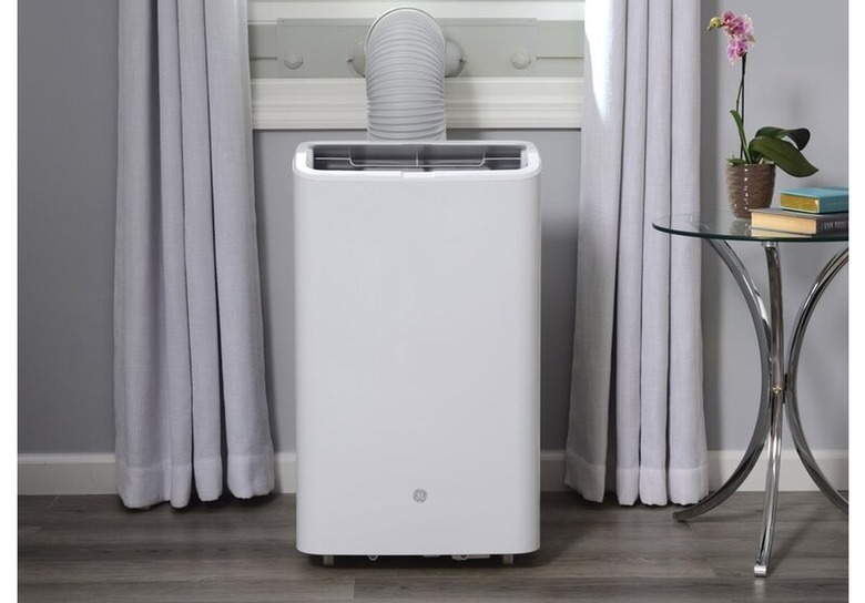 white portable air conditioner; light-gray wall and curtains, white woodwork, and a small glass/metal accent table with books and a pink potted orchid