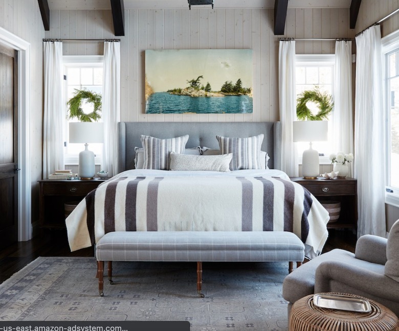 Bedroom with striped quilt, bench, chair, rug, upholstered headboard, pillows, wreaths, curtains.