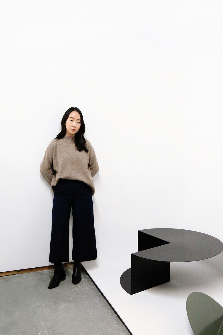 Furniture designer Nina Cho standing, wearing long black pants and a beige sweater with black boots.