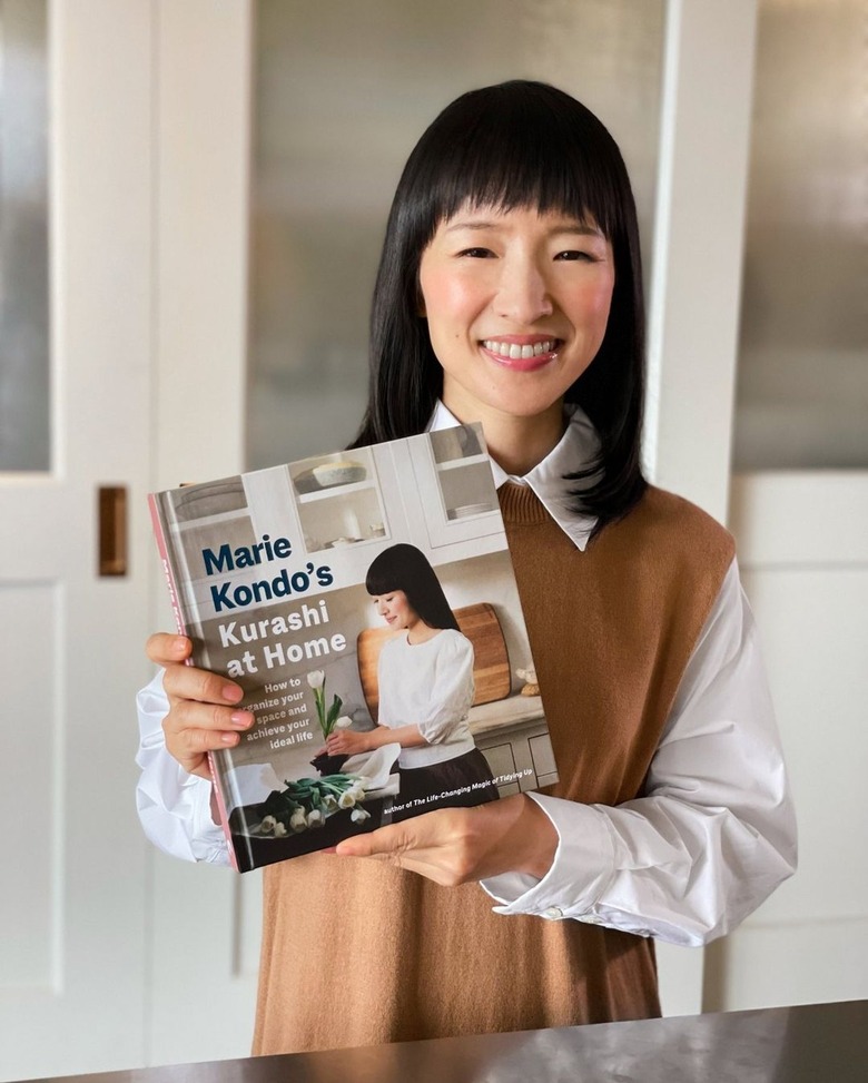 Marie Kondo in a white and brown shirt holding up a book titled 