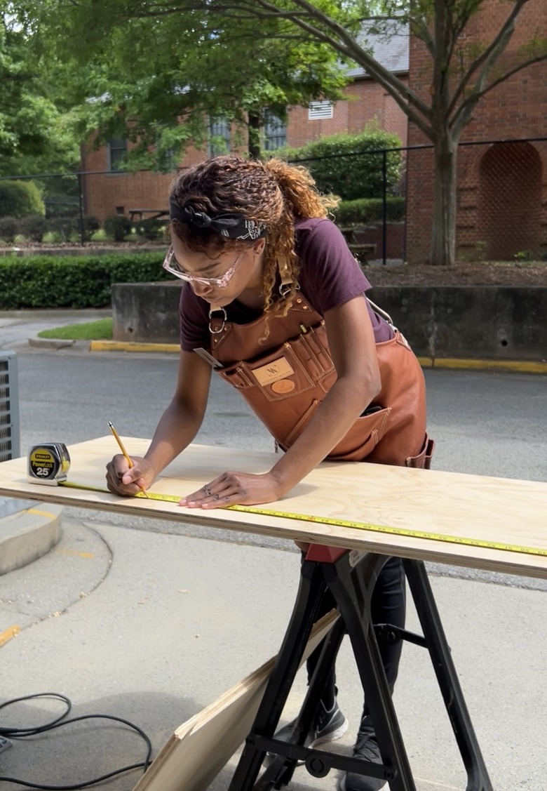 Char Miller-King, a Black person with medium-length curly brown hair, using a pencil to draw on a piece of light wood.