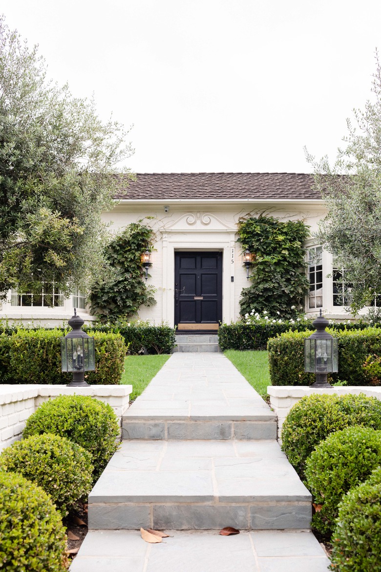 a regal white house with black door, a long concrete path with evenly spaced green hedges that are well manicured