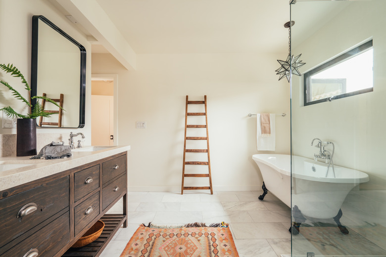 A spacious bathroom with a wood vanity with double sinks, a freestanding clawfoot tub, a glass shower door, a colorful rug, a decorative ladder leaning against the wall