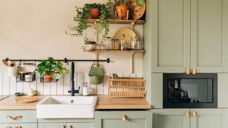 kitchen with light green cabinets, light wood stained countertops, leather hardware pulls, white apron front sink, and black hanging rail, as well as open shelving with plants, bowls, and jars