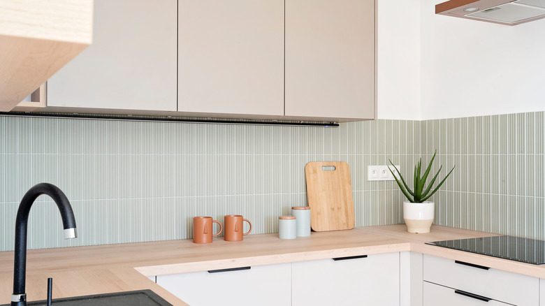 kitchen with while lower cabinets, light wood countertops, beige upper cabinets, grey-green vertical tile backsplash, and black fixtures/hardware