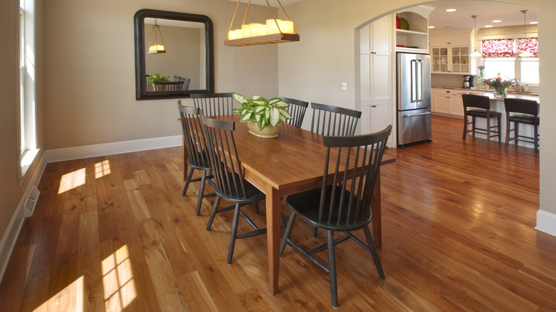 A large dining room feels empty with only a small table and chairs in the center. There is a mirror on the wall and a hanging light fixture over the table. The room is open to the kitchen on one side.