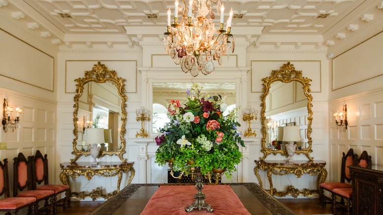 A very formal dining room with tons of architectural details. A large crystal chandelier hangs over an ornate dining table with a tall flower arrangement in the center. There are formal chairs lined against the wall and two ornate gold console tables with mirrors flank the fireplace.