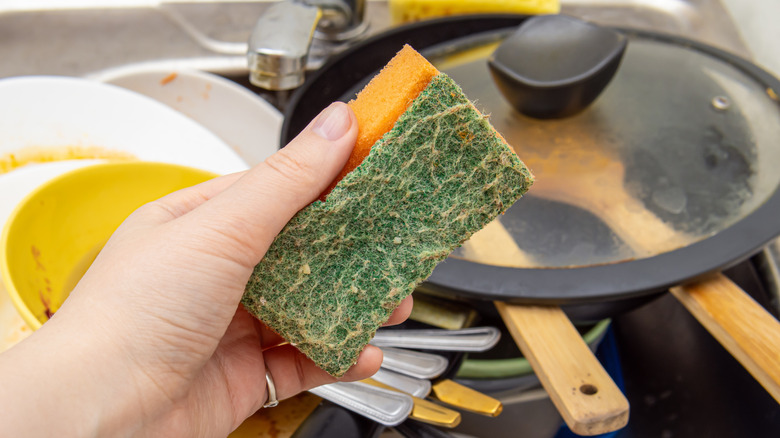 female hand holds dirty sponge with sink with dirty dishes in the background