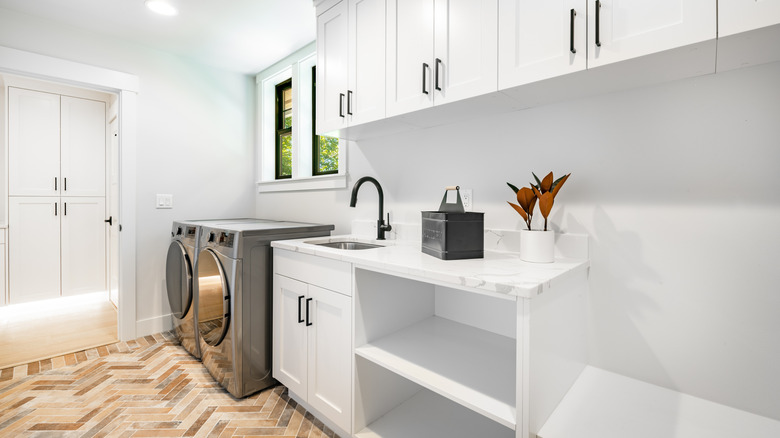 Laundry room with herringbone brick floor, laundry machines under the window, white lower cabinets with open shelves for hamper baskets, and white upper cabinets