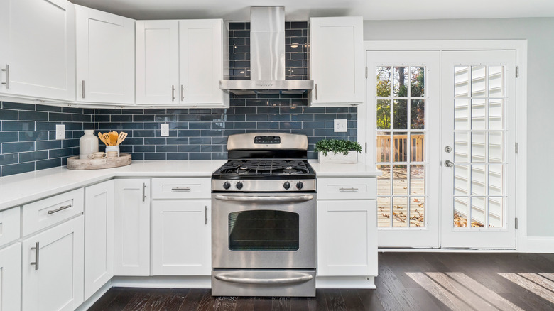 modern kitchen with white shaker cabinets, dark blue subway tile backsplash, stainless steel stove and hood, white countertops, and a pair of white french doors leading to an outdoor deck